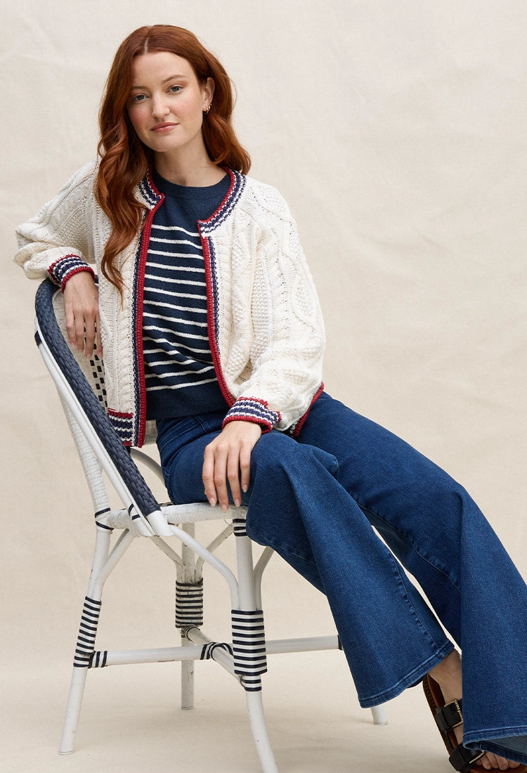 Woman sitting on a chair wearing Maritime Jacket and Pamela Tee, a white jacket, striped shirt, and blue pants in an outdoor setting.