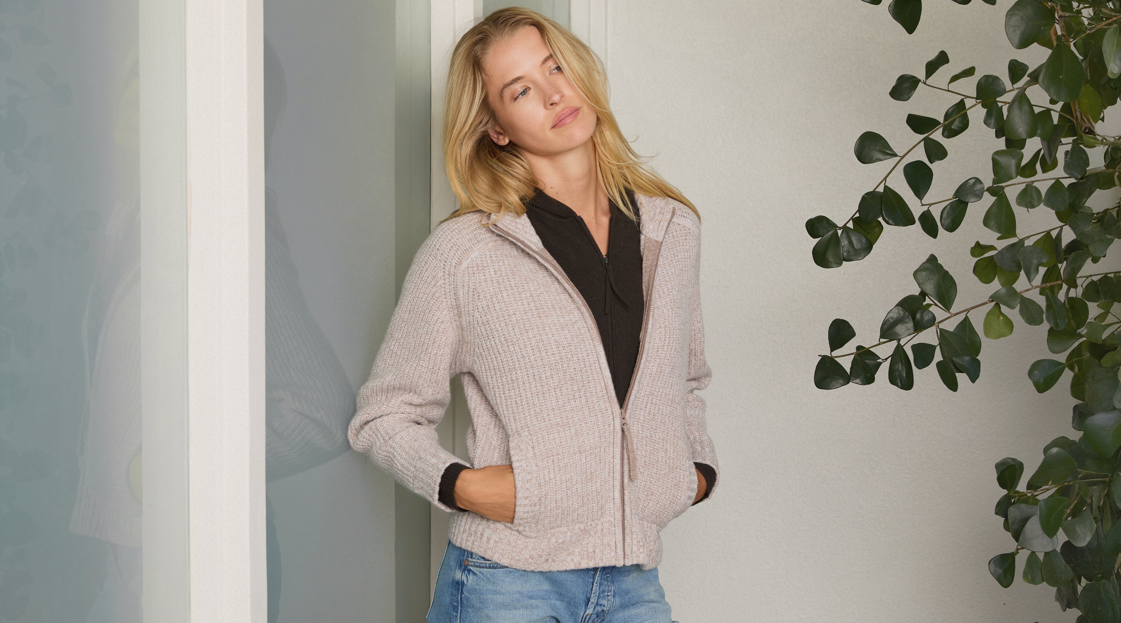 Woman wearing a beige cashmere Tahoe Zip Up sweater by Margaret O'Leary with a plain background and plant on the side.