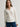 A woman wearing a fitted long-sleeve button-front blouse in off-white, paired with dark blue jeans, photographed against a plain light gray studio background.
