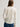 A woman viewed from the back wearing a cream-colored long-sleeve collared shirt and blue jeans against a light gray studio background.