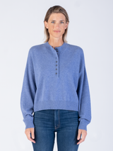 Model wears a cropped blue pullover with a button closure at the front and ribbed details at the neckline, hem and cuffs and a pair of jeans.