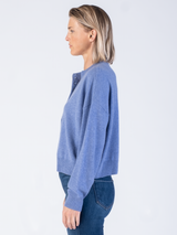 Model wears a cropped blue pullover with a button closure at the front and ribbed details at the neckline, hem and cuffs and a pair of jeans.