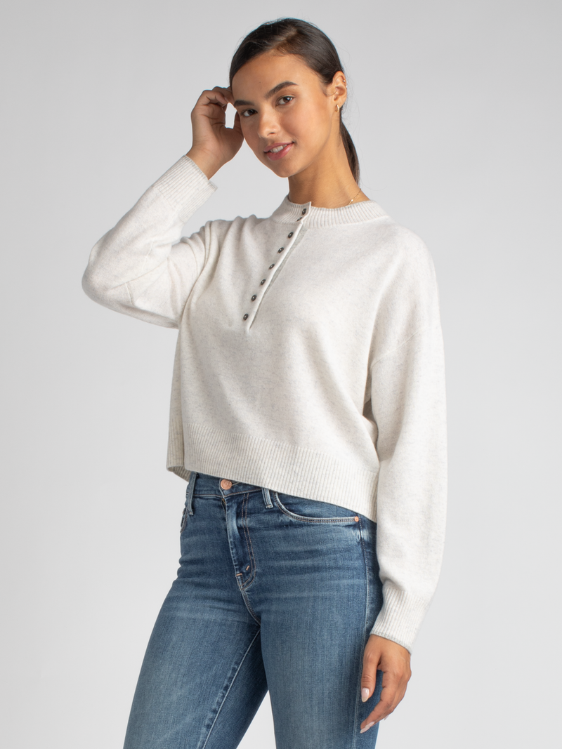 Model wears a cropped white grey pullover with a button closure at the front and ribbed details at the neckline, hem and cuffs and a pair of jeans.