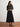 Woman wearing a dark gray sweater and black pleated skirt standing against a white wall.