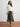 Woman wearing a white jacket and patterned skirt standing on a wooden floor with a white wall background