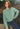 Woman in a Sage cotton boucle Lola Pullover sweater and blue jeans standing in front of plants