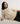 A woman wearing a cream cable-knit sweater with lace-up front detailing and black pants, reclining against a plain light gray studio background.