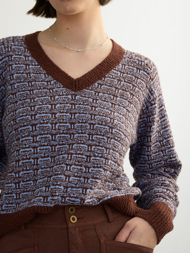 Closeup of a person wearing the Jacquard Vee, a brown and blue patterned sweater by Margaret O'Leary paired with brown pants