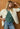 Woman wearing a white Rhys Quilted Jacket over a green Bianca Cashmere Sweater by Margaret O'Leary and blue jeans against a wooden door.