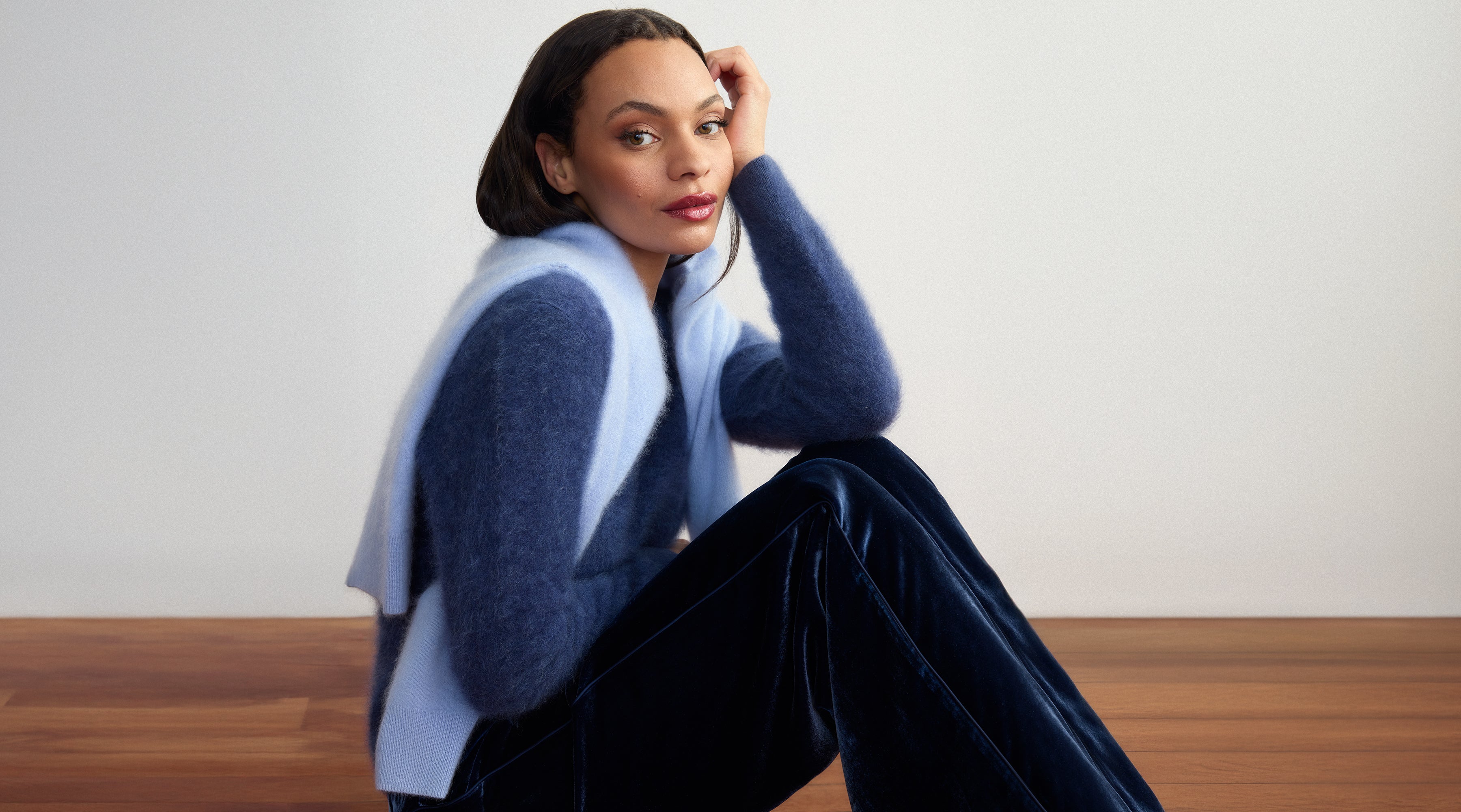 Woman wearing a blue brushed cashmere sweater and dark velvet pants by Margaret O'Leary sitting on a wooden floor.