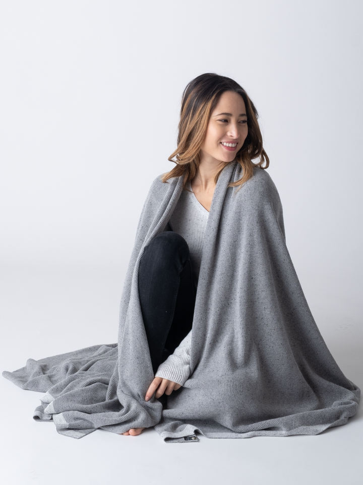 A woman sitting, wearing a large, grey cotton blanket draped over her shoulders.