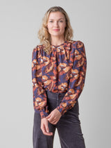 Front view of the model wearing an orange, red and purple floral printed blouse and a pair of grey pants.