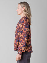 Side view of the model wearing an orange, red and purple floral printed blouse and a pair of grey pants.