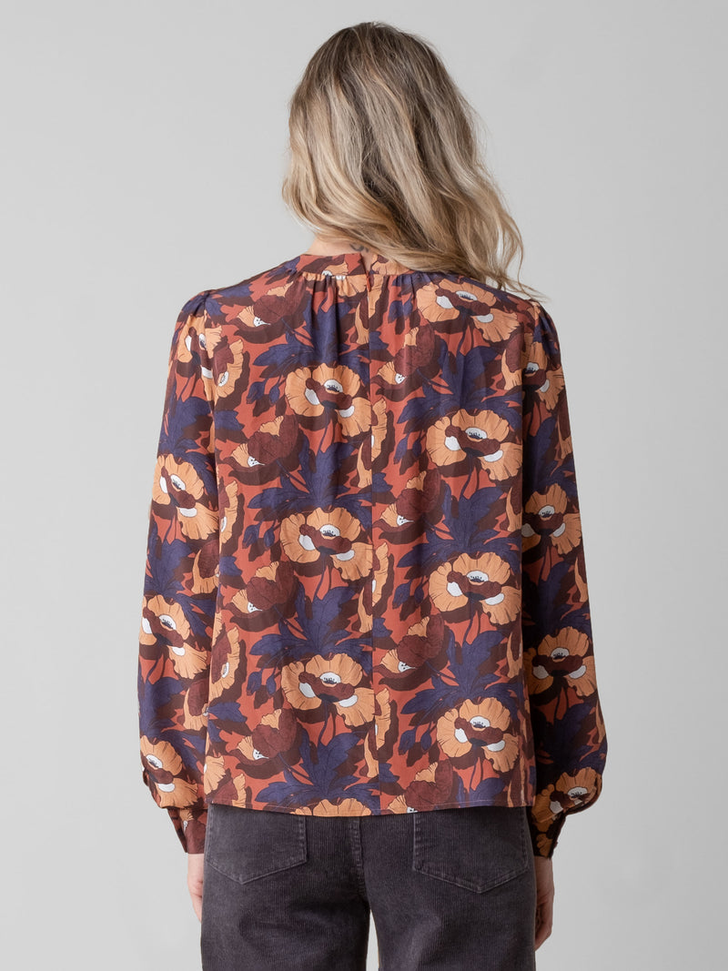 Back view of the model wearing an orange, red and purple floral printed blouse and a pair of grey pants.