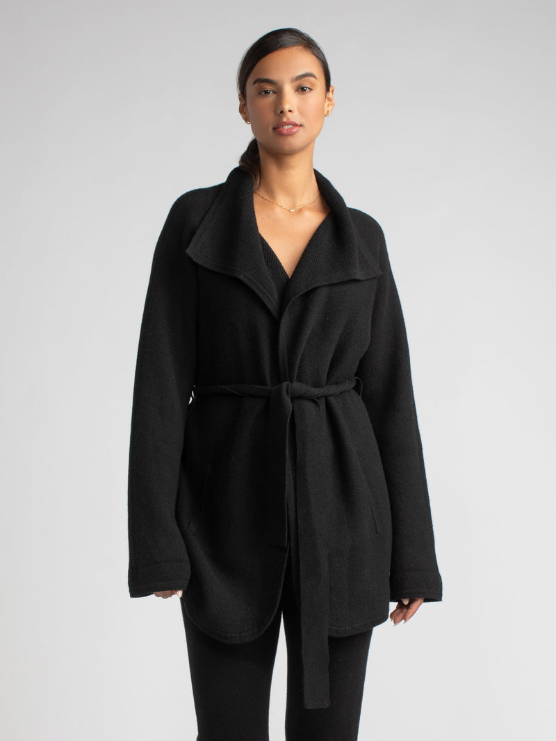 Model wearing a black two pockets jacket with a waist tie and a pair of pants.