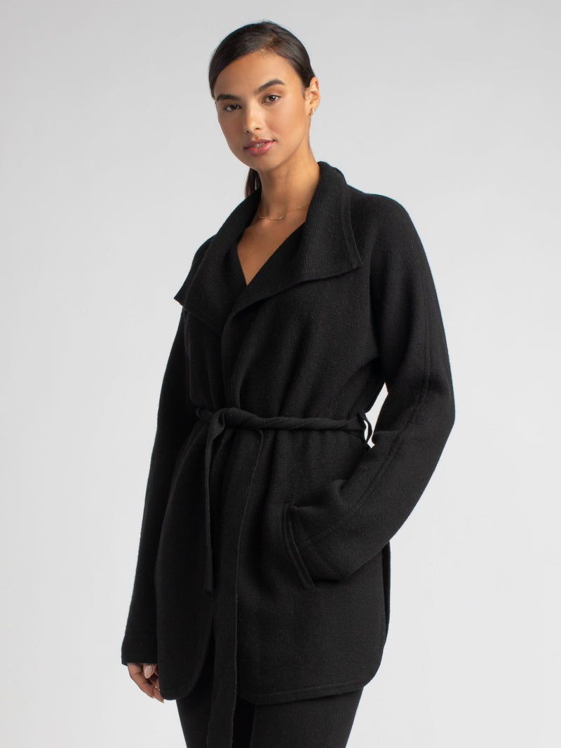 Model wearing a black two pockets jacket with a waist tie and a pair of pants.